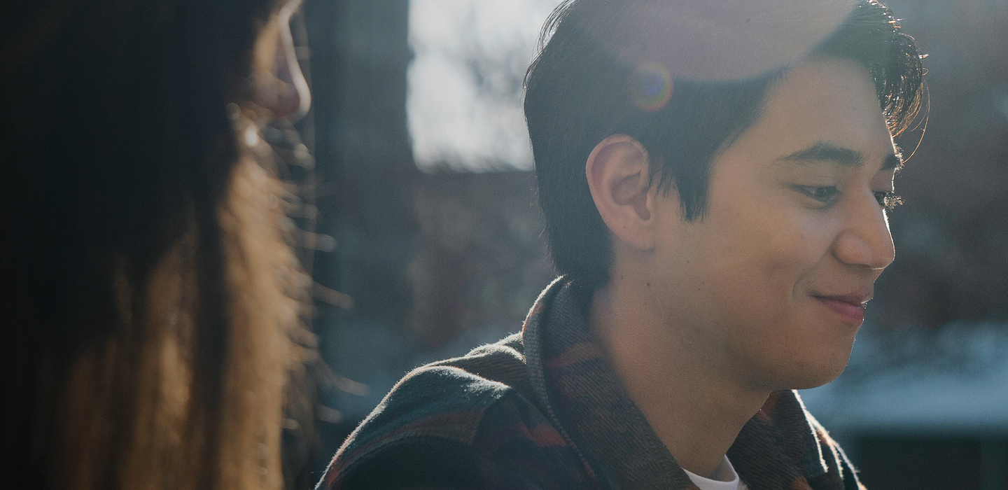 A close-up of a person in profile with short hair, lit by warm sunlight and smiling, with another blurred figure in the foreground.