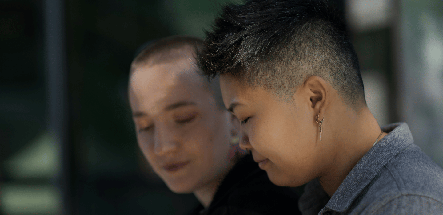 Two people seated close together outdoors, one in focus with short hair and earrings looking downward, while another person appears blurred in the background.