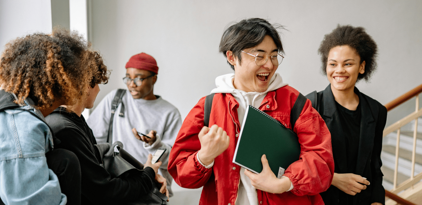 A group of students stand together indoors, one holding a notebook and raising a fist while others smile and talk nearby. 