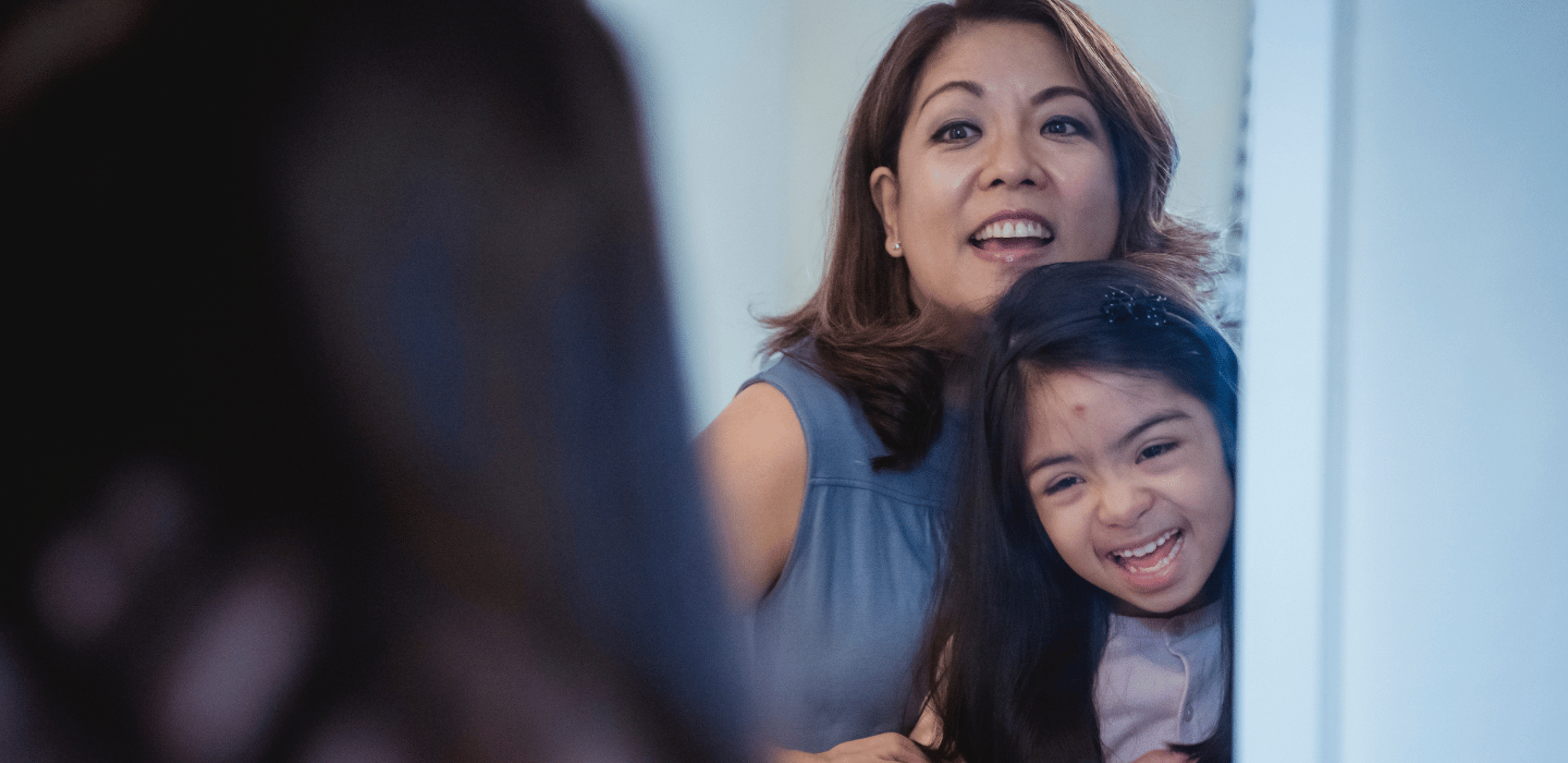 A person holding a young person close while both look toward a mirror, smiling broadly.