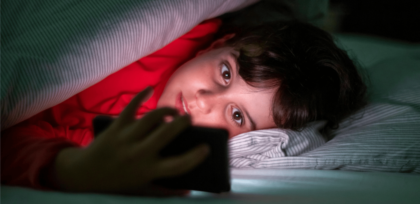 A young person in bed lying under a blanket in dim light, looking at a smartphone that illuminates their face. 