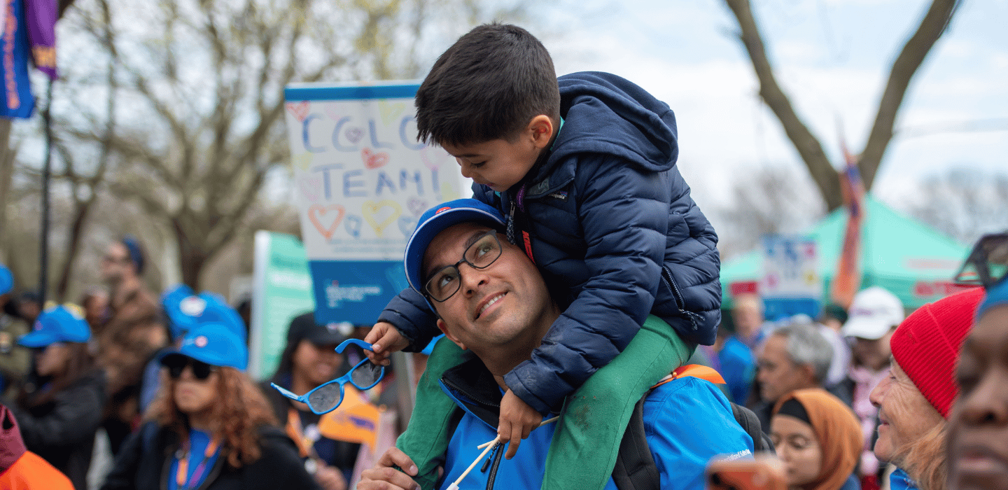 A child sits on an adult’s shoulders and look at each other in a crowd at BMO Walk so Kids Can Talk, while the child is holding sunglasses.