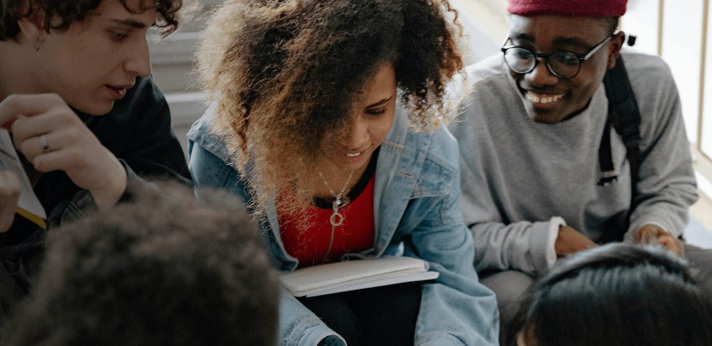 A group of students sitting on steps, looking at a phone together and talking while holding notebooks.