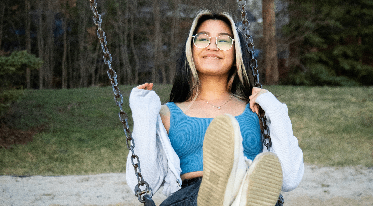 A young person smiling with glasses sits on a swing outdoors, holding the chains while facing the camera, with grassy and wooded areas in the background.