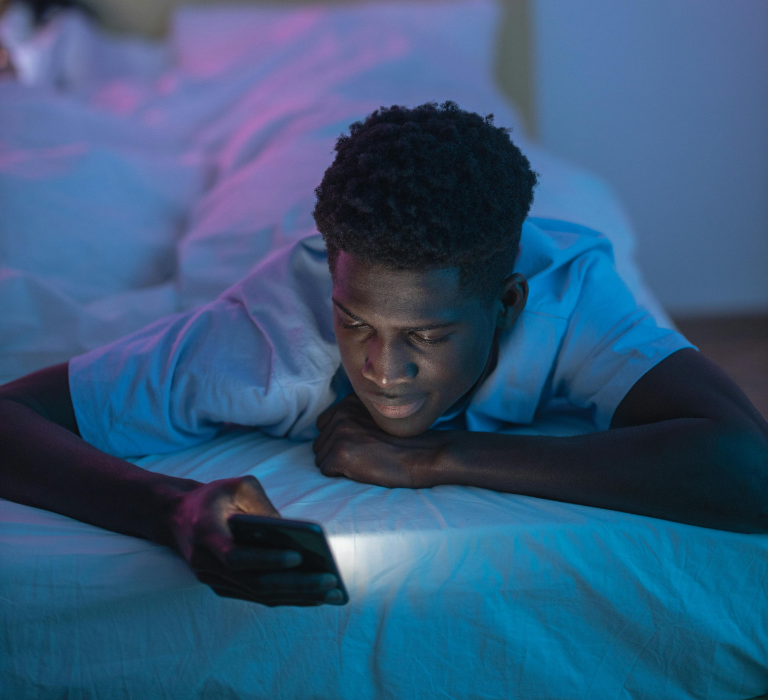 A person lies on a bed looking at a smartphone, with the screen casting a soft glow in a dim room with blue and purple lighting.