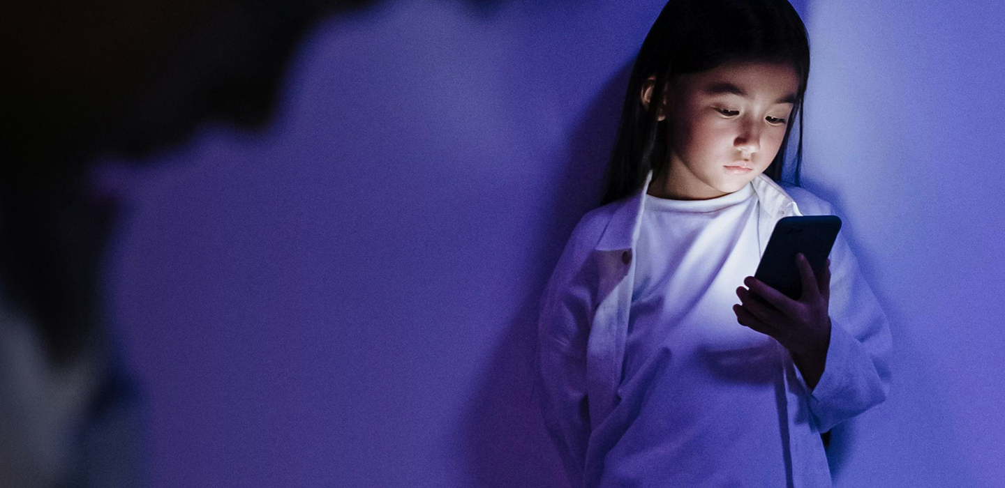 A young person looks at a glowing phone in a dim, blue-lit room.