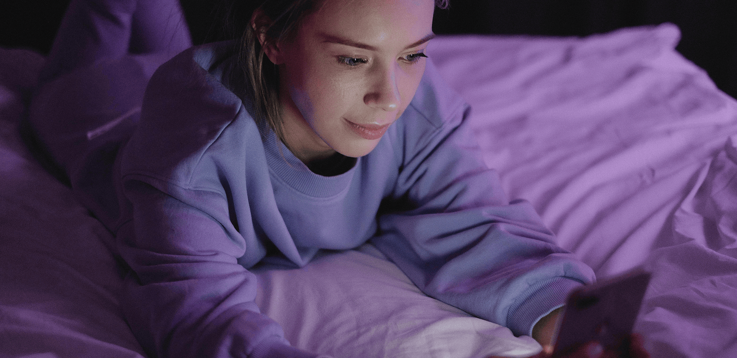 A person lies on a bed using a smartphone, illuminated by soft purple and blue lighting in a dark room.