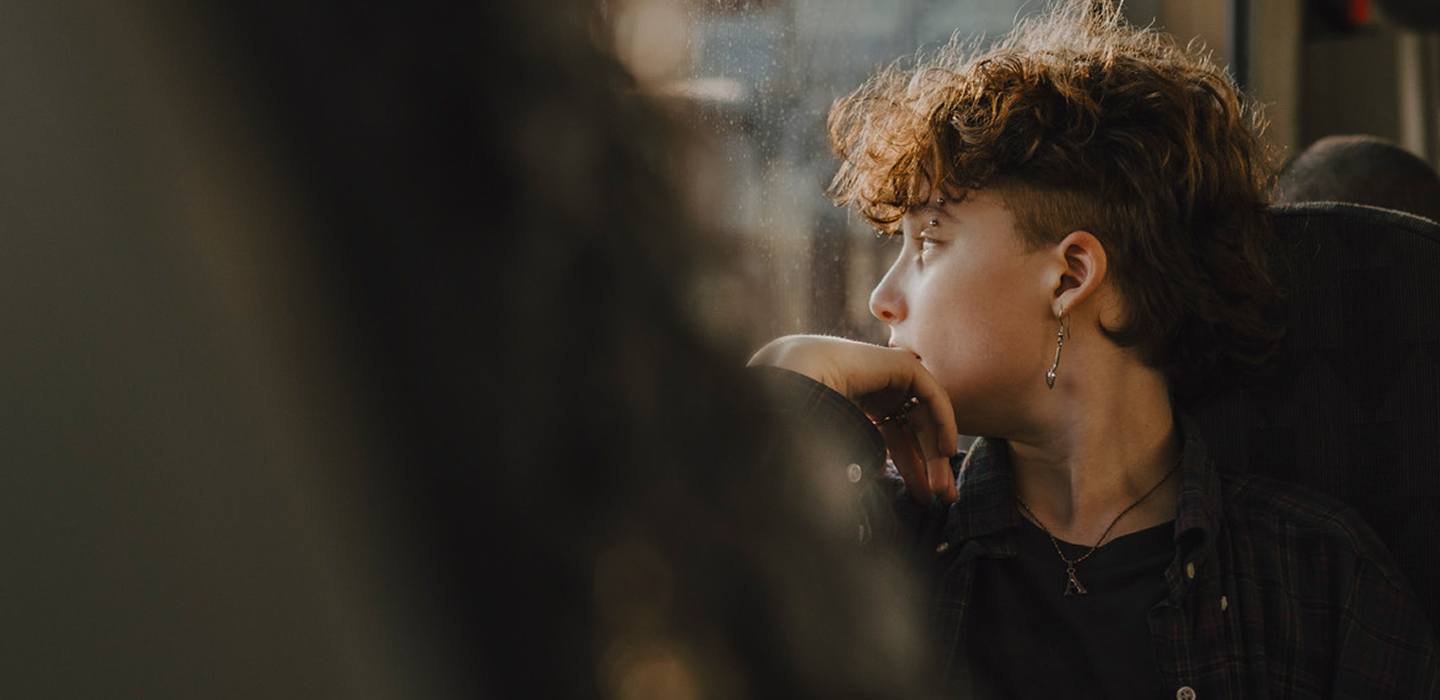 A person with short curly hair and an earring rests their chin on their hand while looking out a window, with soft light highlighting their face.