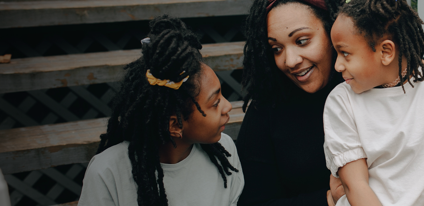 An adult sits with two young people on outdoor steps, leaning in close together while talking with each other. 