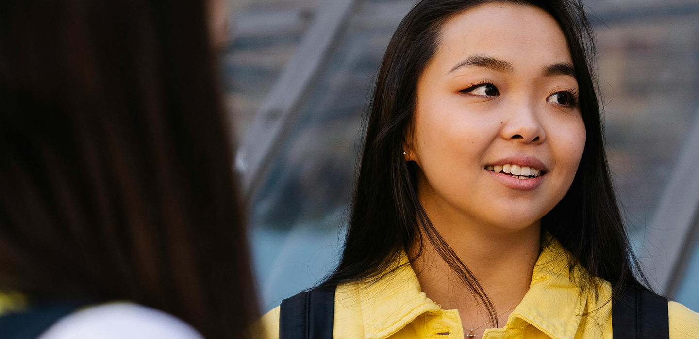 A young person in a yellow jacket and a backpack holds papers while talking with someone outdoors near a glass structure.