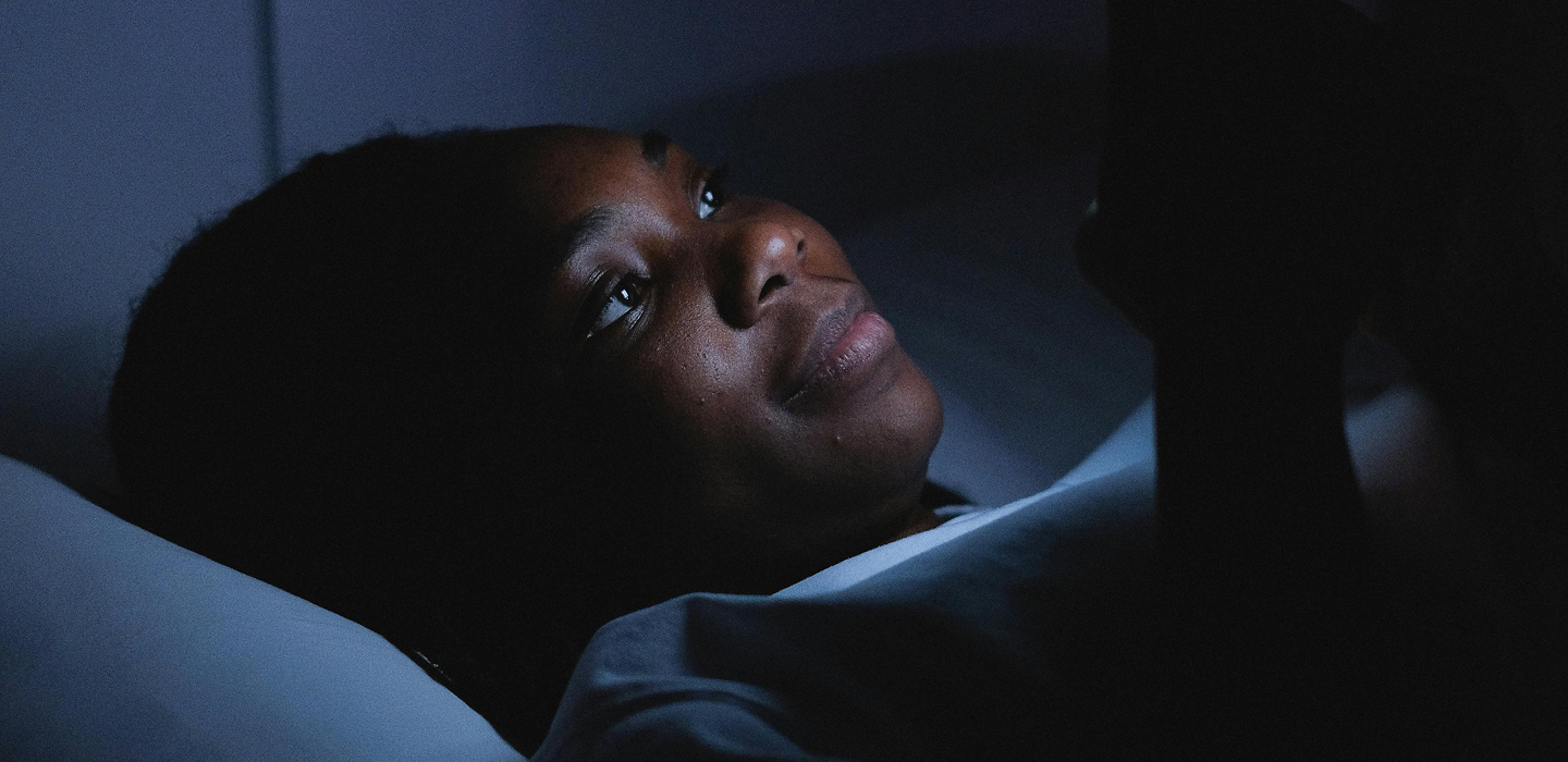 A young person lying in bed in a dark room, illuminated by the light of a smartphone held in one hand.
