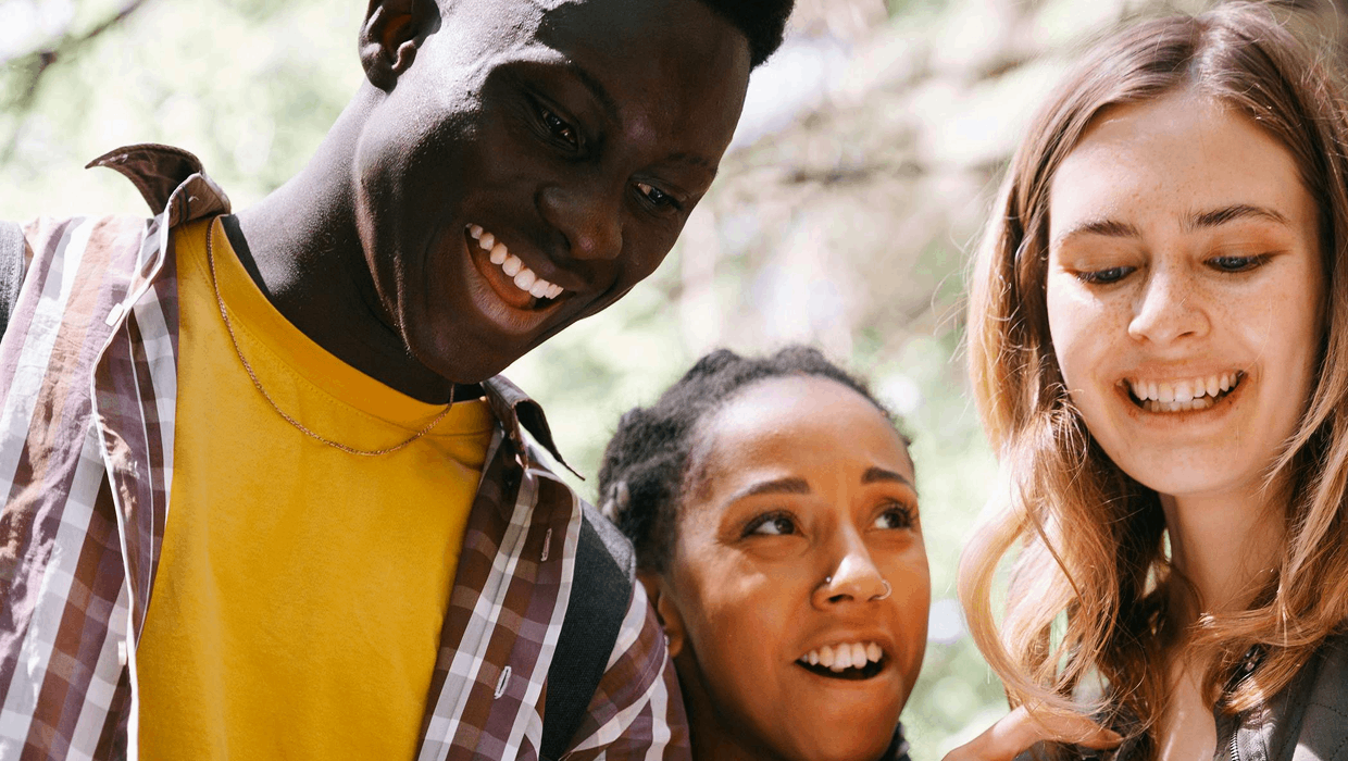 Three people stand close together outdoors, looking down at something and smiling, with soft natural light and greenery in the background. 