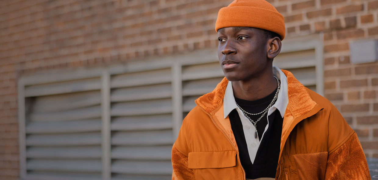 A person wearing an orange hat and jacket stands in front of a brick wall, looking off to the side. 
