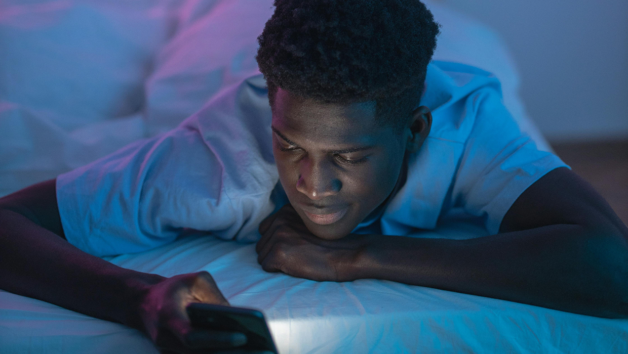 A person lies on a bed looking at a smartphone, with the screen casting a soft glow in a dim room with blue and purple lighting.