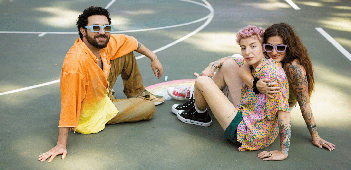 Three people sit on an outdoor court with colorful chalk drawings, wearing casual clothes and sunglasses while relaxing together.