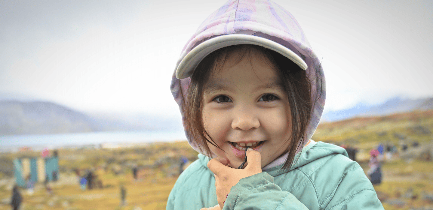 A young person in a cap and jacket standing outdoors, smiling at the camera with a hand near their mouth, and a blurred natural landscape in the background.