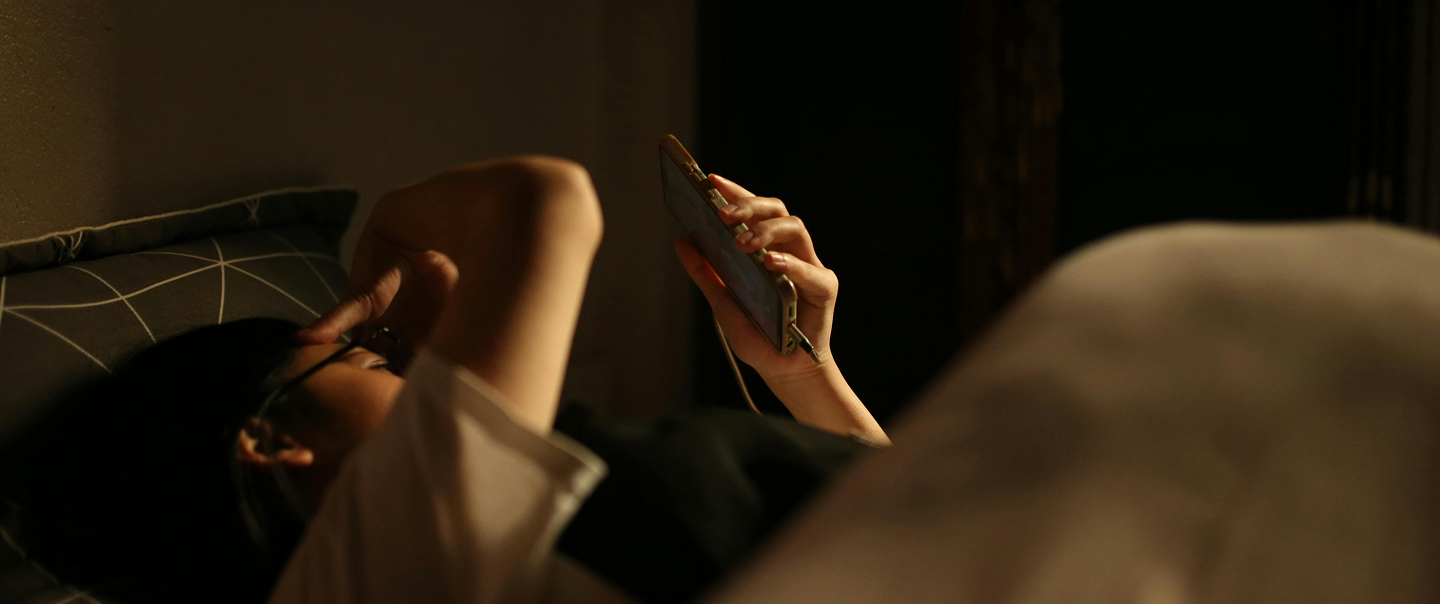 A person lies on a bed in warm, dim lighting, holding a smartphone above their face while another arm rests over their head.