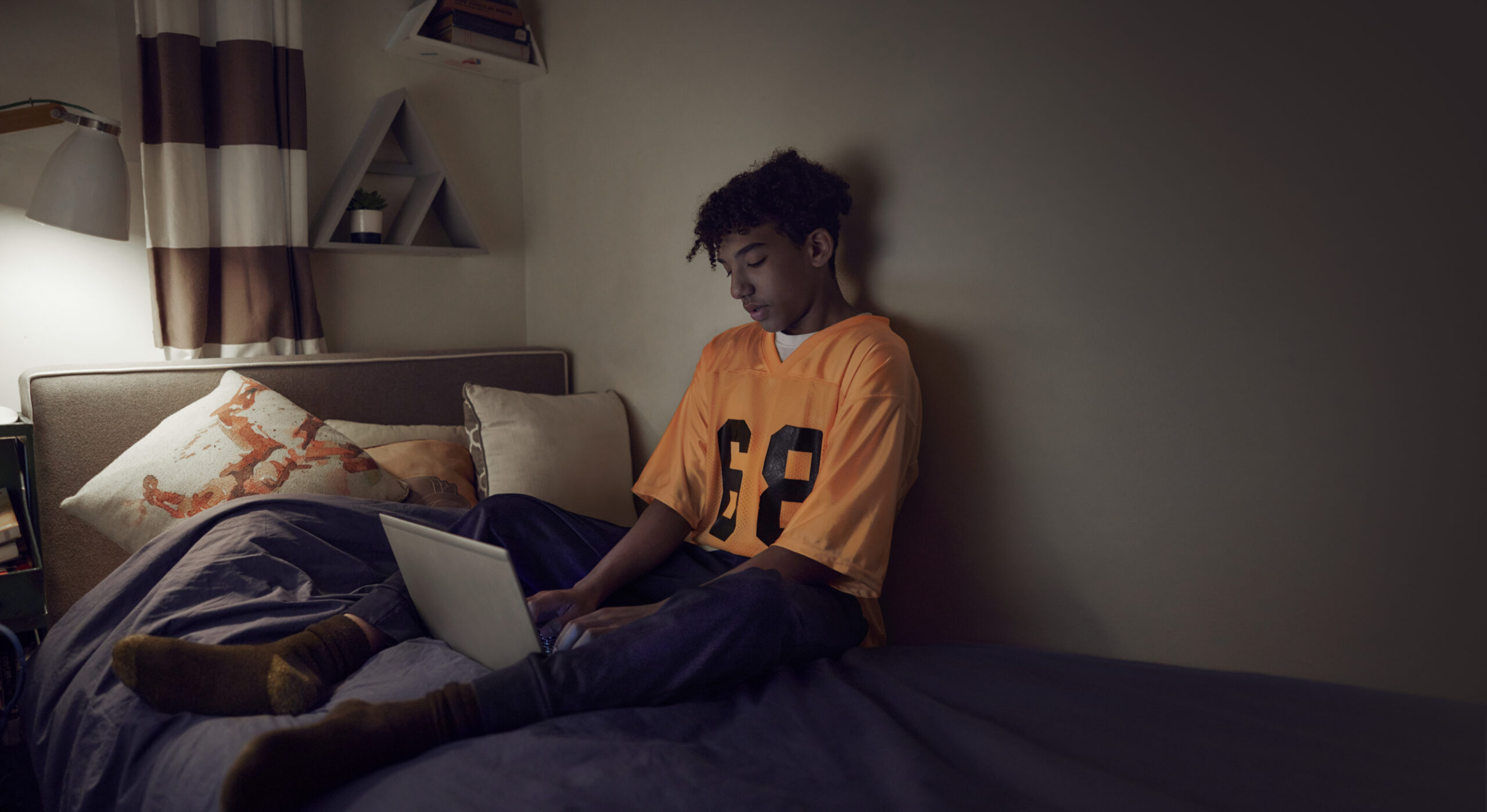 A person sits on a bed using a laptop, with a bedside lamp casting warm light in a dim bedroom.