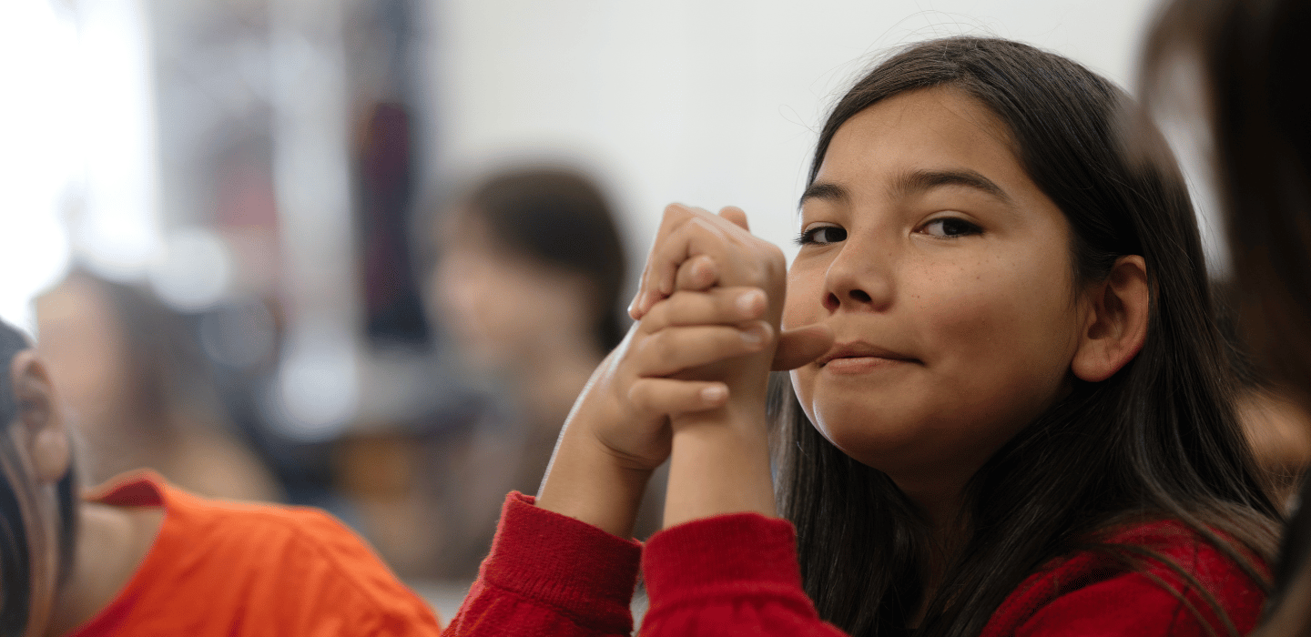A young person in a red sweater sits at a table with hands clasped, looking toward the camera, with other people blurred in the background.