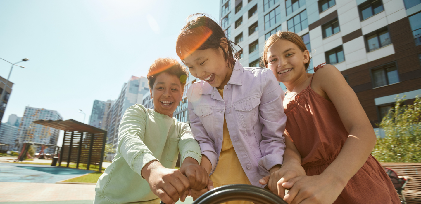 Three young people at a playground smile and hold onto a structure together, with tall buildings and bright sunlight in the background. 