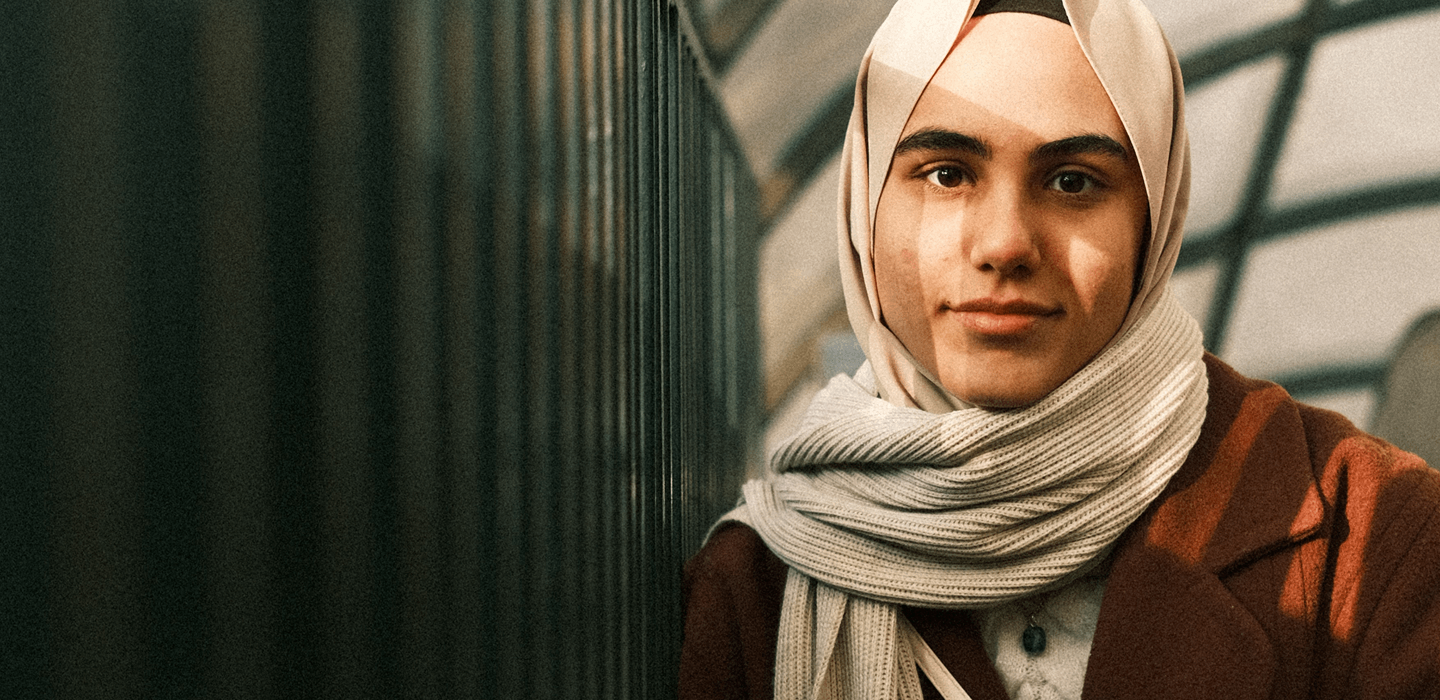 A person wearing a headscarf and long coat sits beside a railing in a covered walkway.