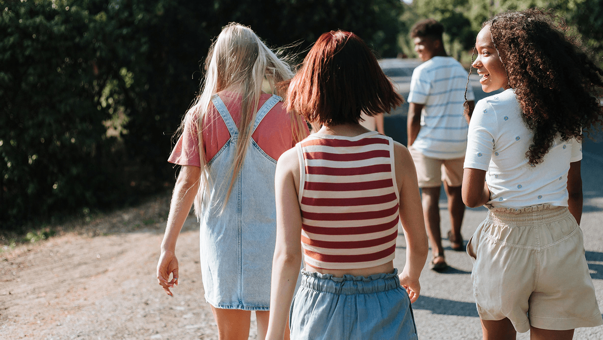 A group of four young people walk along a sunlit path, seen mostly from behind, with trees and greenery lining the road.
