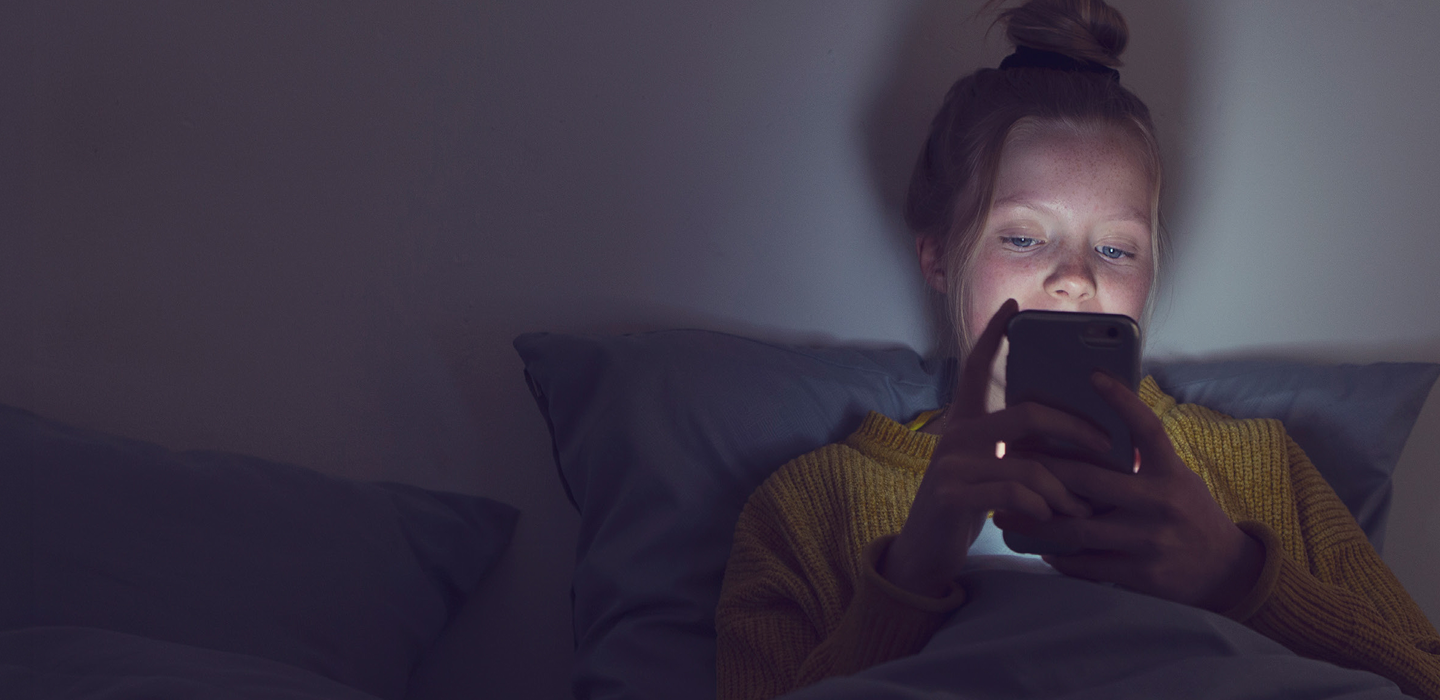 A young person sits in bed under a blanket, using a smartphone that lights their face in a dim room. 