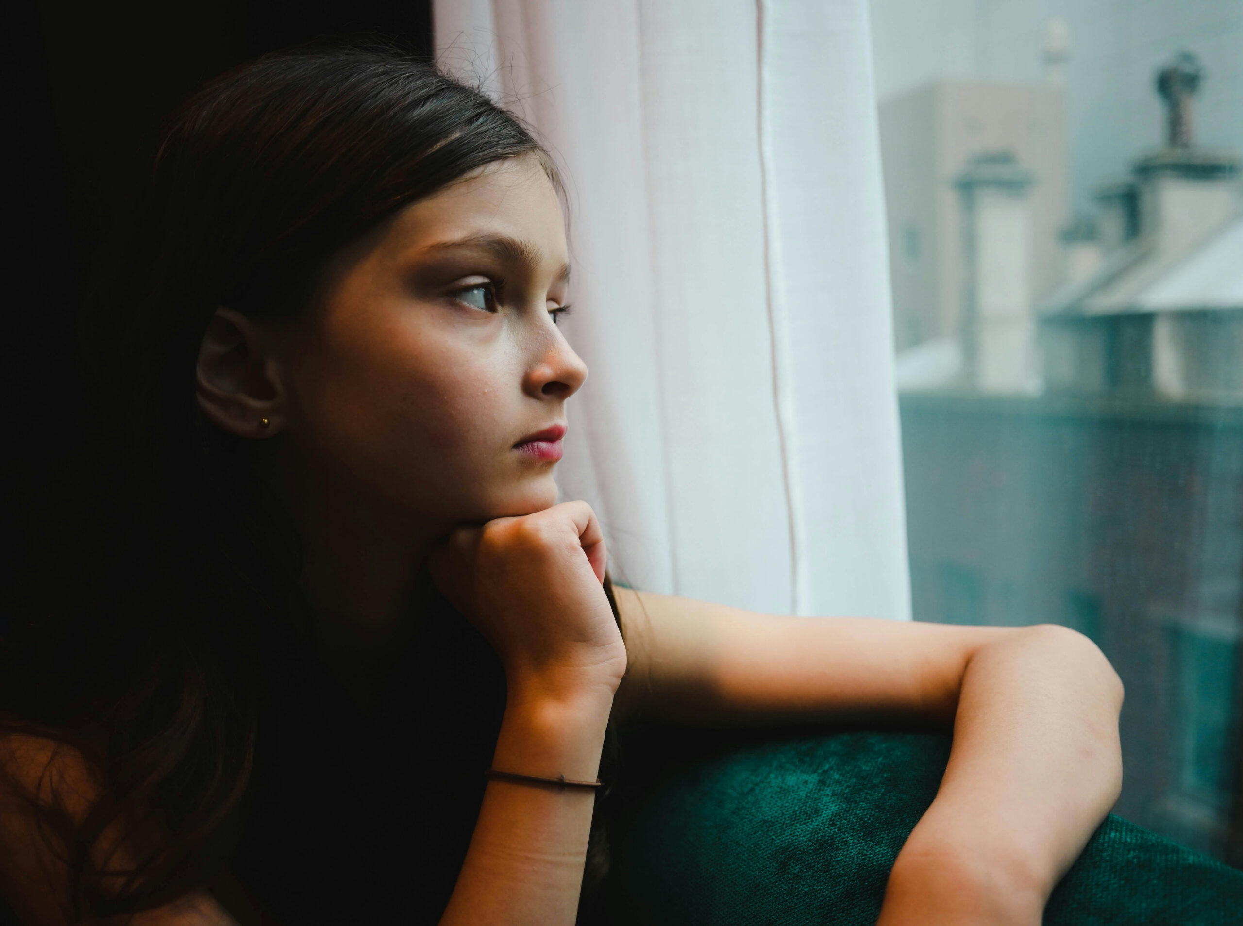 A young person seated indoors beside a window, resting their chin on their hand while looking outward.
