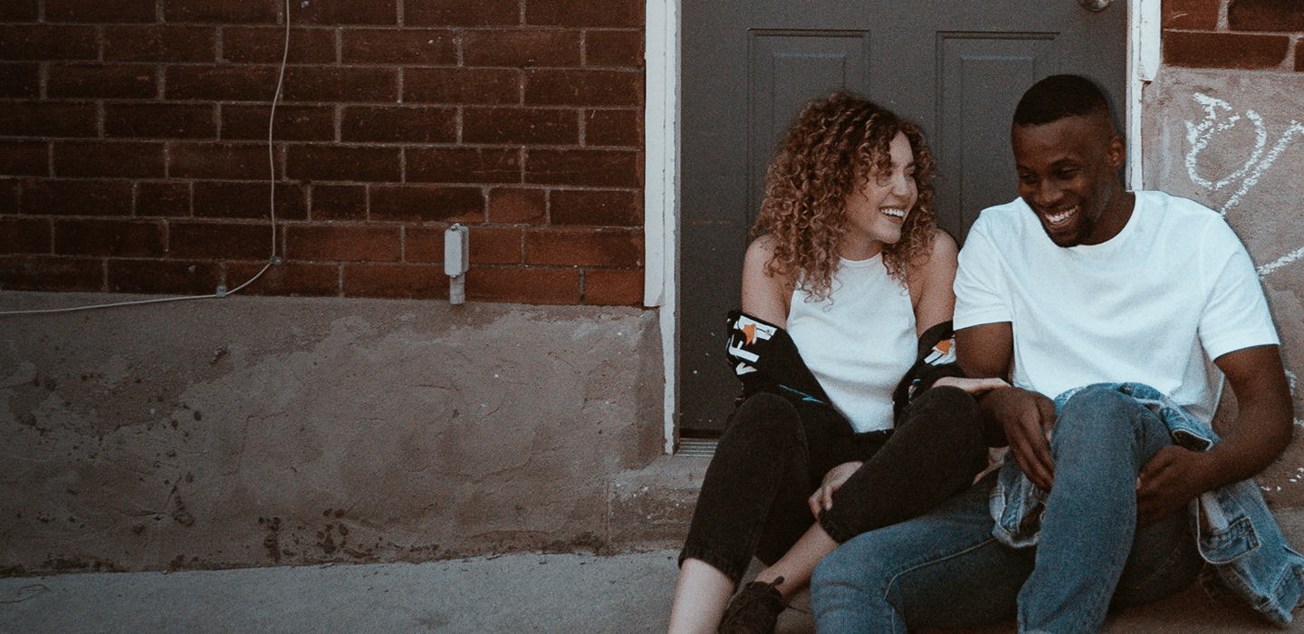 Two people sit on steps outside a brick building, leaning toward each other and laughing. 